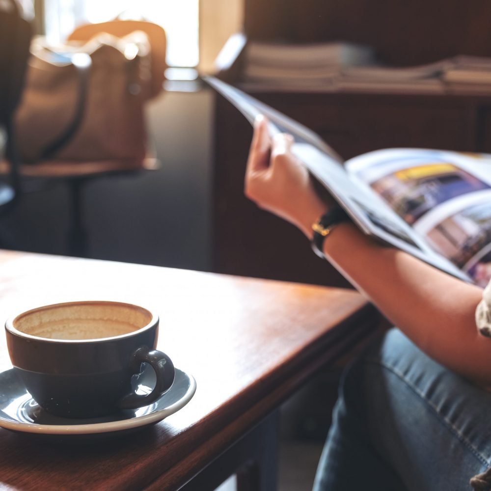 Closeup image of a woman reading a book with coffee cup on wooden table in modern cafe